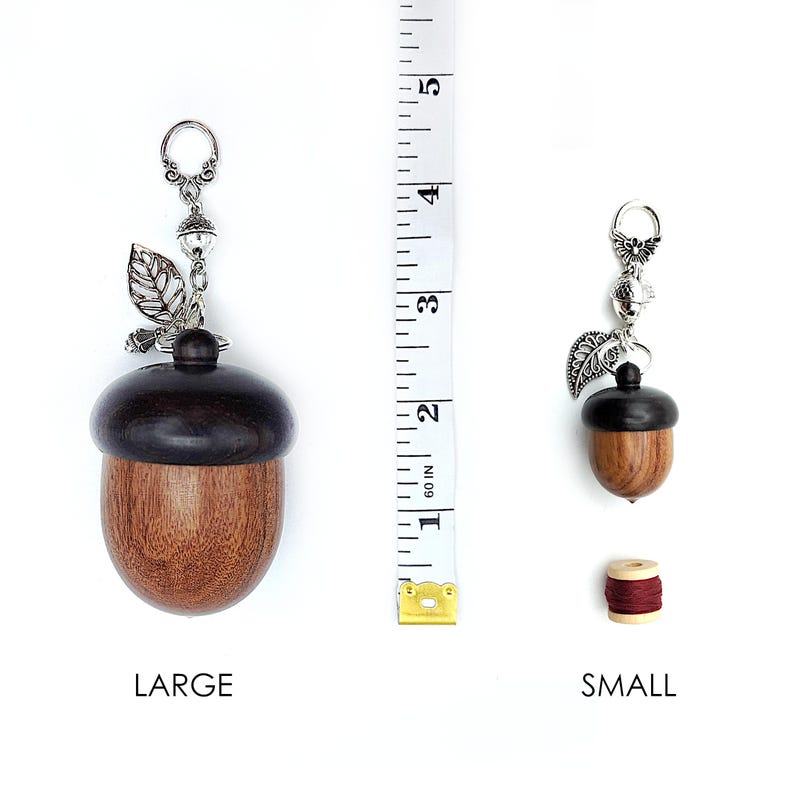 May include: Two wooden acorn-shaped thread spool holders with silver acorn and leaf charms. The larger holder is 2 inches tall and the smaller holder is 1 inch tall. The words "LARGE" and "SMALL" are printed below each holder.