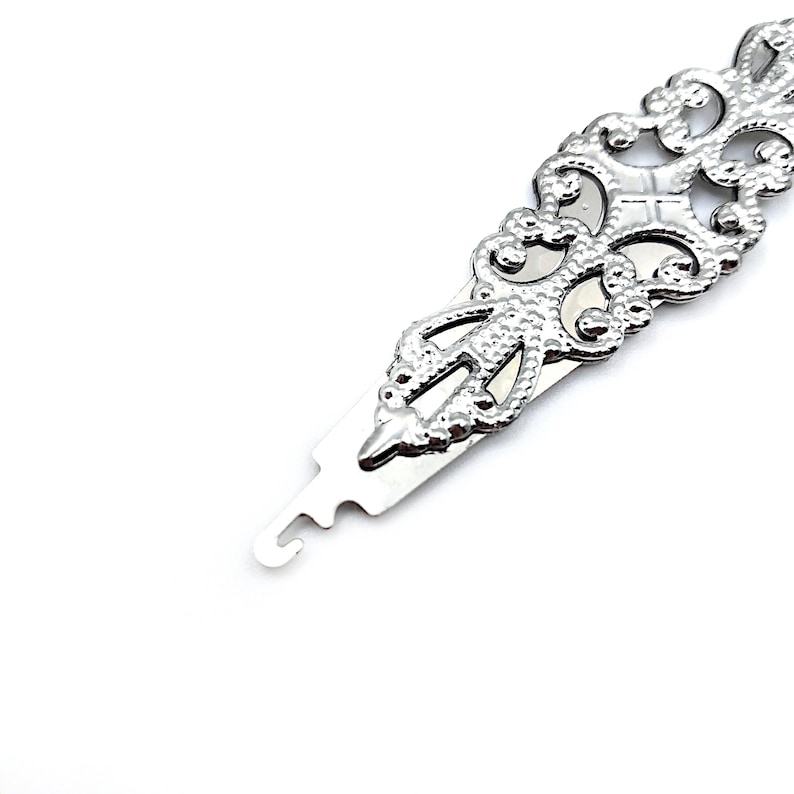 May include: A silver metal hook tool with a decorative, ornate design. The tool has a small loop at one end and a pointed end at the other.