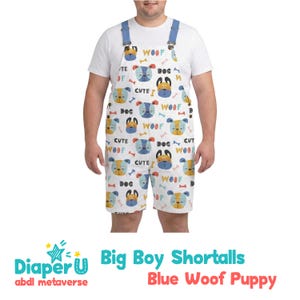 Op de afbeelding: Witte korte tuinbroek met een blauw bandontwerp, met een herhalend patroon van cartoonhondengezichten, botten en de woorden "Woof", "Dog" en "Cute". De tekst "Big Boy Shortalls Blue Woof Puppy" staat onderaan.