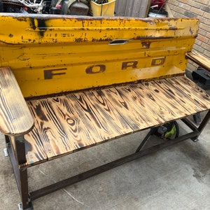 May include: A yellow metal bench with a wooden seat and armrests. The bench is made from a repurposed Ford truck tailgate. The tailgate has the word "FORD" embossed on it.