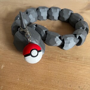 Puede incluir: Un brazalete gris flexible segmentado con un clip plateado y un llavero Poké Ball rojo y blanco.