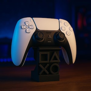 Op de afbeelding: Een witte en zwarte draadloze gamecontroller op een zwarte standaard. De controller heeft een witte bovenkant en een zwarte onderkant met knoppen en joysticks. De standaard toont de PlayStation-symbolen.