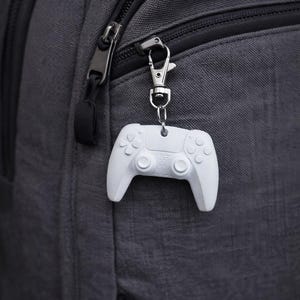 Op de afbeelding: Een witte controller sleutelhanger hangt aan een zilveren clip die aan een grijze rugzak is bevestigd. De sleutelhanger is een miniatuur replica van een game controller, met knoppen en directionele pads. De rugzak heeft een rits en een zwarte band.