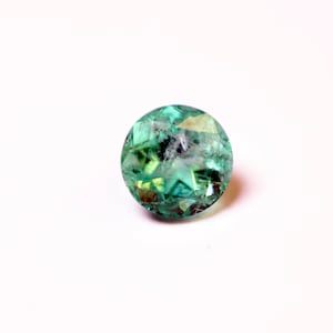 Puede incluir: Una piedra preciosa redonda y facetada de color verde con un tono verde claro. La piedra preciosa está tallada en un patrón de estrella.