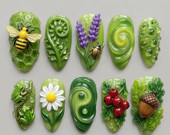 Könnte beinhalten: Ein Set aus zehn dekorativen Nagelkunststücken mit naturinspirierten Motiven, darunter eine Biene auf einer Wabe, ein Farn, Lavendel mit einem Käfer, ein grüner Wirbel, Wassertropfen, zwei Frösche, ein Gänseblümchen, ein Yin-Yang-Symbol, rote Beeren und eine Eichel.