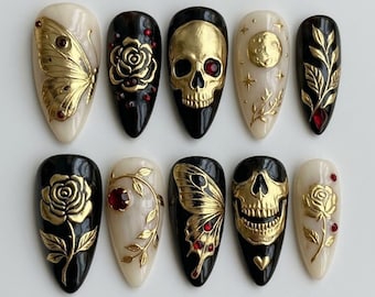 Könnte beinhalten: Ein Set aus zehn mandelförmigen Press-on-Nägeln in einer schwarz-cremefarbenen Farbpalette. Die Designs umfassen goldene, geprägte Totenköpfe, Schmetterlinge, Rosen, Mondphasen und Blattreben, akzentuiert mit kleinen, dunkelroten, runden Strasssteinen.
