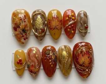 Könnte beinhalten: Ein Set aus zehn künstlichen Fingernägeln mit herbstlichen Motiven. Die Nägel zeigen warme Farben wie Orange, Gelb und Braun mit gemalten Ahornblättern, roten Beeren, Blattgold-Akzenten und wirbelnden Marmormustern in glänzender Optik.