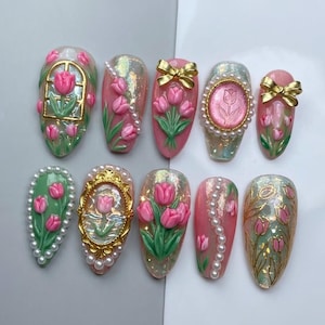 Peut inclure: Un ensemble de dix faux ongles décorés de tulipes roses, d'accents métalliques dorés et de perles blanches. Les ongles présentent diverses textures, notamment des finitions irisées, des cadres dorés et des nœuds sur une base rose et verte.