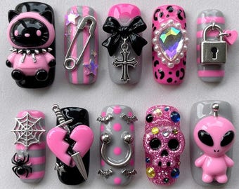 Könnte beinhalten: Ein Set aus zehn künstlichen Fingernägeln in Pink, Schwarz und Grau. Die Verzierungen umfassen eine 3D-Katzenfigur, eine Sicherheitsnadel, eine schwarze Schleife mit Kreuzanhänger, einen schillernden Edelstein, ein Vorhängeschloss, ein Spinnennetz und einen Totenkopf.