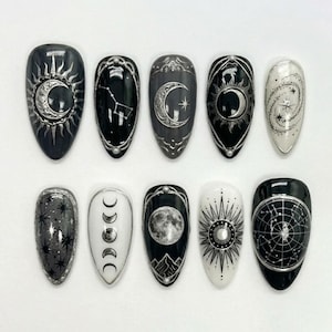 May include: A set of ten almond-shaped press-on nails featuring celestial designs in black, white, and silver. The patterns include crescent moons, suns, stars, constellations, lunar phases, and a galaxy motif with intricate metallic silver line work.
