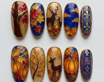 Könnte beinhalten: Ein Set aus zehn ovalen Zierplaketten mit farbenfrohen Designs im Emaille-Stil. Die Motive zeigen herbstliche Bäume, Ahornblätter, Hirsche, Kürbisse und goldene Lilien-Symbole auf blauem und violettem Hintergrund, eingefasst in Goldmetall.