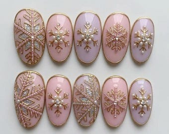 Könnte beinhalten: Ein Set aus zehn ovalen Press-on-Nägeln in Hellrosa mit goldenen und silbernen Schneeflockenmotiven. Einige Nägel zeigen große, komplexe Schneeflockenmuster mit Glitzer, während andere mit kleinen, zarten goldenen Schneeflocken und Perlen verziert sind.