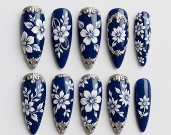 Könnte beinhalten: Ein Set aus zehn mandelförmigen Press-on-Nägeln in tiefem Marineblau. Jeder Nagel ist mit erhabenen weißen Blumenmustern, filigranen silberfarbenen Metallverzierungen an den Spitzen oder Basen und kleinen klaren Strasssteinen versehen.