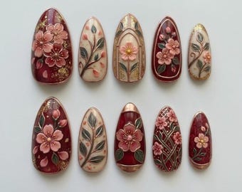 Könnte beinhalten: Ein Set aus zehn mandelförmigen Press-on-Nägeln in einer Farbpalette aus tiefem Burgunderrot und Creme. Die Designs zeigen erhabene, rosafarbene 3D-Kirschblüten, grüne Blätter, Blattgoldakzente und filigrane Gittermuster mit goldenen Rändern.