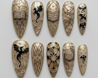 May include: A set of ten almond-shaped press-on nails featuring a cream base with black and gold accents. Designs include black dragons, open books with text, reaching hands, geometric star patterns, and clouds against a starry night sky.