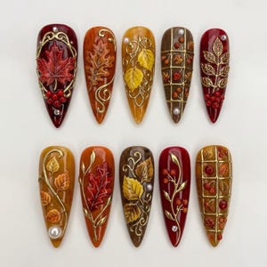 May include: Ten almond-shaped press-on nails featuring autumn-themed designs. The set includes deep red, orange, yellow, and brown bases adorned with raised gold metallic leaf motifs, berries, grid patterns, and small white pearl accents.
