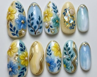Könnte beinhalten: Ein Set aus zehn ovalen künstlichen Fingernägeln in den Farben Hellblau, Creme und Gelb. Die Designs umfassen 3D-Blumenmotive, handgemalte blaue Blätter, Blattgoldakzente, kleine weiße Perlen und eingearbeitete Muschelfragmente.