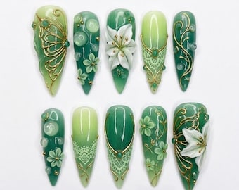 Könnte beinhalten: Ein Set aus zehn stilettoförmigen künstlichen Fingernägeln in Grüntönen. Die Designs zeigen weiße 3D-Blüten, goldene Filigranmuster, durchscheinende grüne Kugeln und filigrane, spitzenartige weiße Details auf einer glänzenden, grünen Basis.