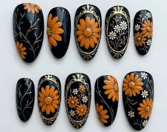 Könnte beinhalten: Ein Set aus zehn mandelförmigen Press-on-Nägeln mit glänzend schwarzer Basis. Die Designs zeigen erhabene orangefarbene Gänseblümchen, zarte goldene Ranken und filigrane goldene Rahmen um Gruppen von orangefarbenen und weißen Blumenmustern.