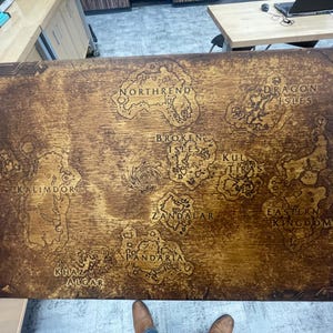 May include: A wooden map with a detailed, engraved design. The map features various landmasses labeled with names like "Kalimdor," "Northrend," and "Eastern Kingdoms." The wood has a warm, brown tone, and the map is held by a person.