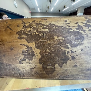 Puede incluir: Un mapa de madera rectangular grande con un diseño de mapa del mundo grabado detallado. El mapa presenta masas terrestres, océanos e ilustraciones estilizadas de dragones, barcos y otros elementos de fantasía. La madera tiene un tono marrón cálido.