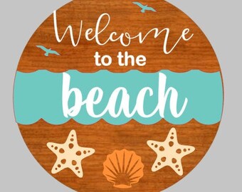 May include: Round wooden sign with the text "Welcome to the beach" in white cursive lettering. The background is brown wood with a blue wave design. There are two white starfish and a seashell in orange and white.