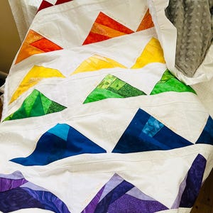 May include: A white quilt with a rainbow pattern of triangles. Each triangle is made of different fabrics and colors. The quilt is folded in half and has a ruffled edge.