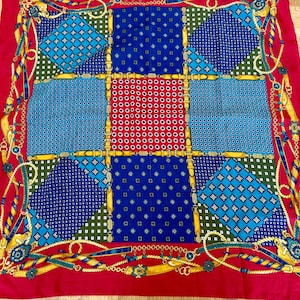 May include: A square scarf with a patchwork design. The scarf features a red border and a central pattern of blue, green, and red squares with various geometric and dotted patterns. Gold-colored rope and belt details adorn the edges.
