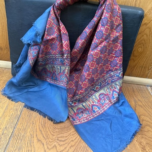 May include: A vintage scarf with a vibrant pattern in red, blue, and gold. The scarf features a detailed geometric design and is draped over a black leather case. The scarf has a solid blue fringe.