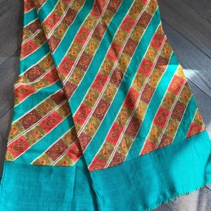 Puede incluir: Una bufanda textil rectangular con rayas diagonales en color verde azulado vibrante que alternan con bandas de cuadrados rojos y marrones. Cada cuadrado contiene un patrón de cachemira dorado. La bufanda tiene un borde liso y flecos.