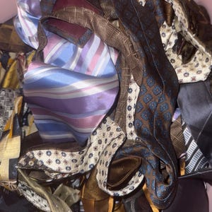 Könnte beinhalten: Eine Sammlung von Krawatten mit verschiedenen Mustern und Farben, darunter eine auffällige hellblau-violett gestreifte Krawatte, eine braune Krawatte mit blauem geometrischem Muster, eine cremefarbene Krawatte mit kleinen dunklen Punkten und weitere.