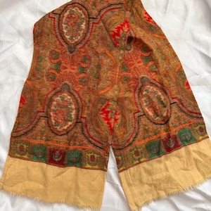 Puede incluir: Una bufanda rectangular con un complejo estampado de cachemira en tonos naranja quemado, rojo intenso y verde bosque. El diseño incluye grandes motivos ovalados y un borde decorativo sobre un dobladillo beige claro con flecos.