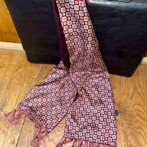 May include: A burgundy scarf with a repeating geometric pattern in white, blue, and gold. The scarf has a fringed edge and is draped over a black leather briefcase. The scarf is made of a soft, silky material.