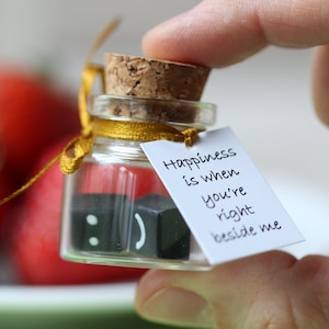 Könnte beinhalten: Ein kleines Glasgefäß mit einem Korkverschluss und einem gelben Band, das darum gebunden ist. Das Gefäß enthält zwei kleine schwarze Blöcke mit Smiley-Gesichtern. Ein weißes Etikett ist an dem Gefäß befestigt mit dem Text "Glück ist, wenn du an meiner Seite bist".