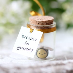 May include: A small glass jar with a cork top and a gold ribbon tied around it. Inside the jar is a small, yellow bee figurine. A white card attached to the jar reads "Bee-lieve in yourself".