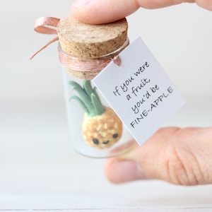 May include: A small glass jar with a cork lid contains a miniature pineapple figurine. A white tag attached to the jar reads "If you were a fruit, you'd be FINE-APPLE".