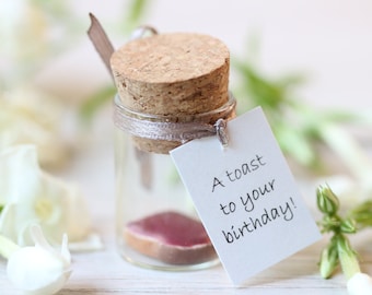 Peut inclure: Un petit bocal en verre avec un bouchon en liège et une tranche de pain grillé à l'intérieur. Une carte blanche attachée au bocal indique "A toast to your birthday!"