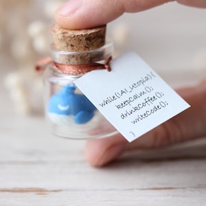 Può includere: Un piccolo barattolo di vetro con un tappo di sughero contiene una statuetta di balena blu. Un'etichetta bianca attaccata al barattolo recita "while(!Al_utopia) { keepcalm(); drinkcoffee(); writeCode(); }"