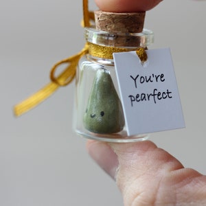 May include: A small glass jar with a cork lid and a gold ribbon tied around the neck. Inside the jar is a green pear-shaped figurine with a happy face. A white tag is attached to the ribbon with the text "You're pearfect".