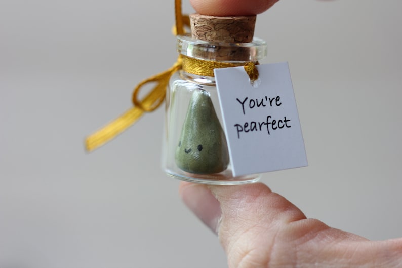 May include: A small glass jar with a cork lid and a gold ribbon tied around the neck. Inside the jar is a green pear-shaped figurine with a happy face. A white tag is attached to the ribbon with the text "You're pearfect".
