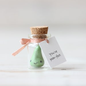 May include: A small glass jar with a cork lid, tied with a pink ribbon, contains a green pear-shaped figurine with a smiling face. A white tag attached to the jar reads "You're pear-fect."