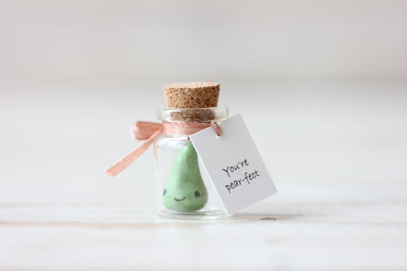 May include: A small glass jar with a cork lid, tied with a pink ribbon, contains a green pear-shaped figurine with a smiling face. A white tag attached to the jar reads "You're pear-fect."