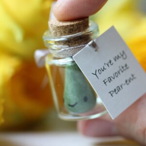 May include: A small glass jar with a cork lid contains a green pear-shaped figurine with a smiling face. A white tag attached to the jar reads "You're my favorite Pear-ent".