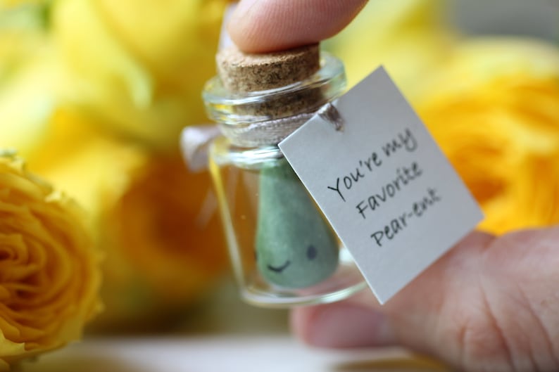 May include: A small glass jar with a cork lid contains a green pear-shaped figurine with a smiling face. A white tag attached to the jar reads "You're my favorite Pear-ent".
