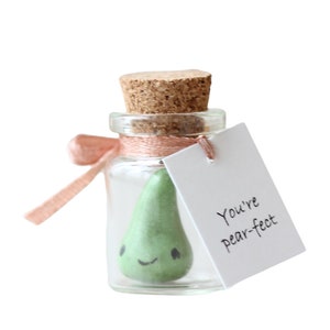 May include: A small glass jar with a cork lid contains a green pear-shaped figurine with a smiling face. A pink ribbon is tied around the jar and a white tag is attached with the text "You're pear-fect".