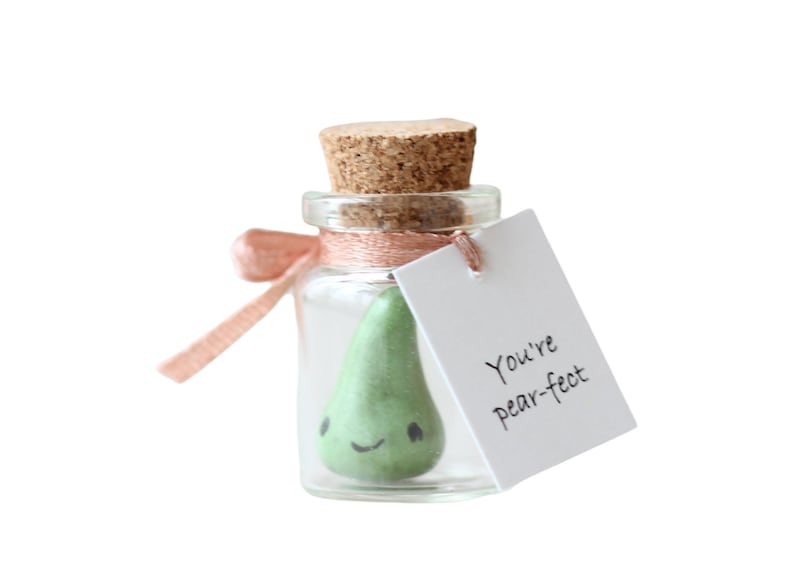 May include: A small glass jar with a cork lid contains a green pear-shaped figurine with a smiling face. A pink ribbon is tied around the jar and a white tag is attached with the text "You're pear-fect".