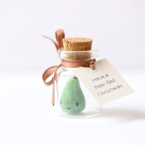 May include: A small glass jar with a cork lid contains a green pear-shaped ornament with a smiling face. A brown ribbon is tied around the jar with a tag that reads "Have a pear-fect Christmas".