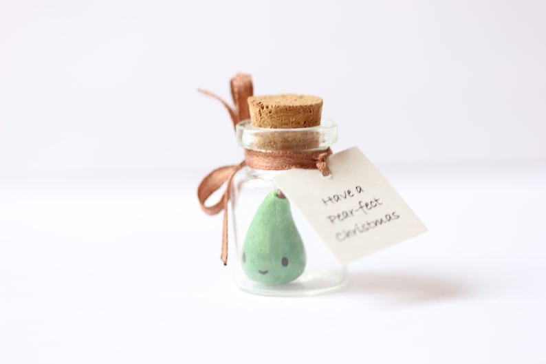May include: A small glass jar with a cork lid contains a green pear-shaped ornament with a smiling face. A brown ribbon is tied around the jar with a tag that reads "Have a pear-fect Christmas".