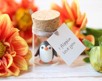 May include: A small glass jar with a cork lid contains a small penguin figurine. The jar has a white tag attached to it with the text "I flippin love you". The jar is surrounded by orange flowers.