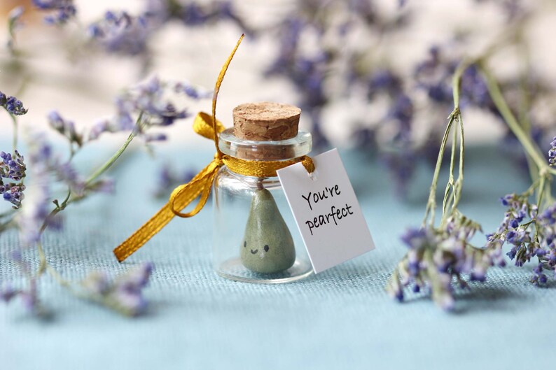 May include: A small glass jar with a cork lid and a yellow ribbon tied around it. Inside the jar is a small green pear with a happy face. A white tag is attached to the ribbon with the text "You're Pearfect".